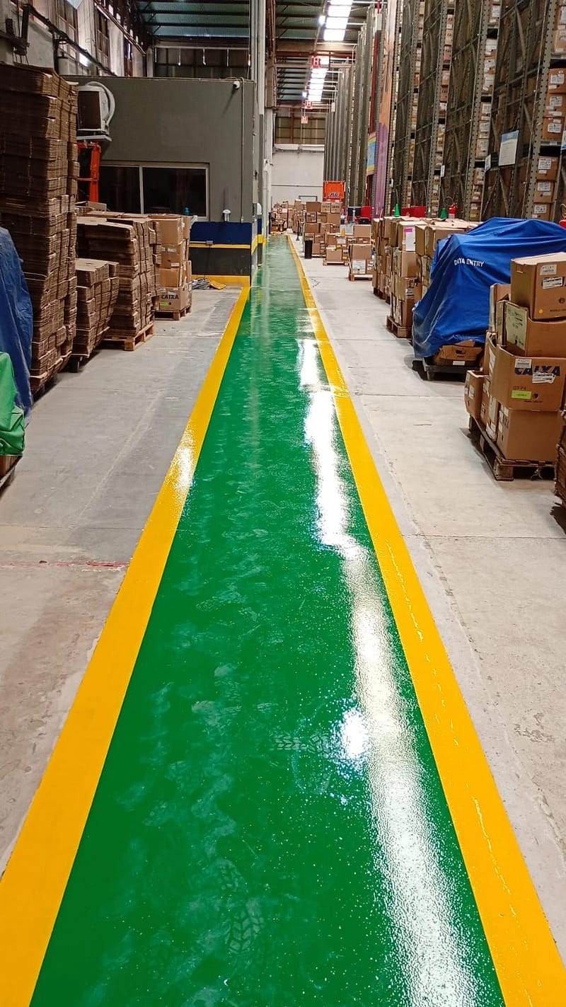 Faixa de pedestres em pintura epóxi verde com bordas amarelas, entre pilhas de caixas em armazém
