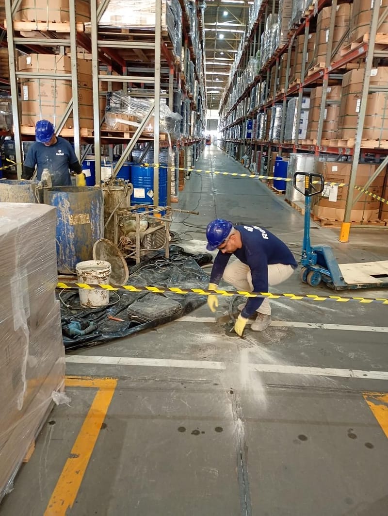 Vista do corredor de um centro de distribuição durante a injeção de calda de cimento no piso, com bomba, misturador e operador junto ao furo