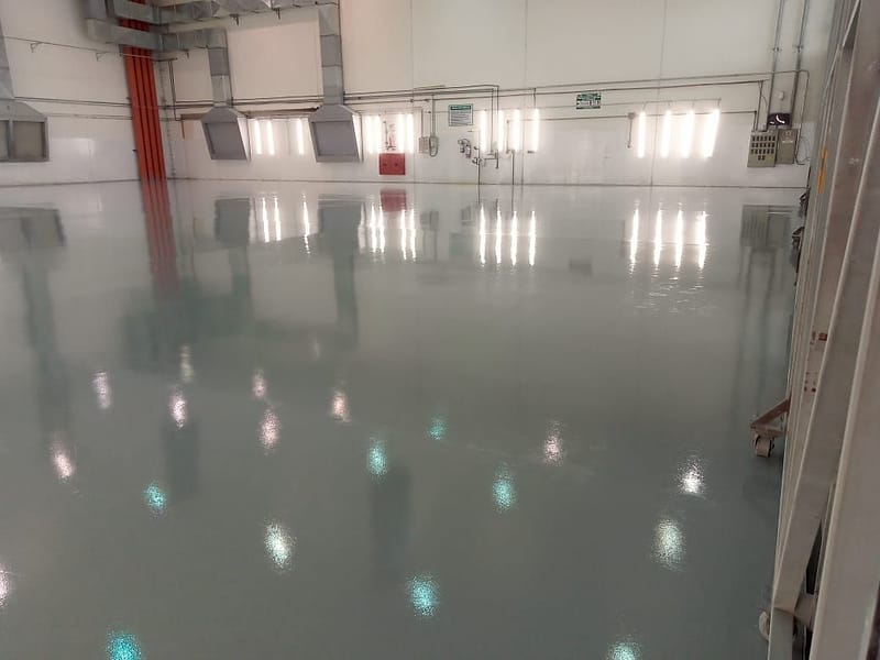 Piso epóxi autonivelante cinza de alto brilho em ampla área de produção, refletindo luminárias e dutos de exaustão