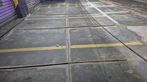 Estacionamento frontal de loja com placas de concreto escurecidas e juntas abertas, isolado por corrente à noite